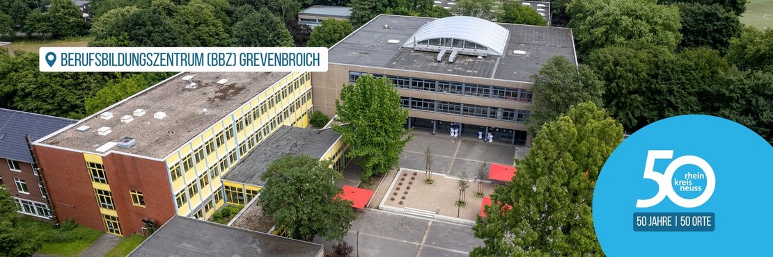 Das Bild zeigt eine Luftaufnahme des Berufsbildunszentrums Grevenbroich.  Man sieht mehrere Gebäude, einen Innenhof mit Bäumen und Sitzgelegenheiten, sowie einen Teil der umliegenden Landschaft. Im Vordergrund befindet sich ein Logo, das auf das 50-jährige Jubiläum des Rhein-Kreises Neuss hinweist.