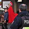 Das Bild zeigt im Vordergrund den Rücken eines Polizisten in Uniform, der auf einen Umzug oder eine Parade blickt.  Im Hintergrund ist ein Mann in römischer Rüstung und einem roten Umhang auf einem Pferd zu sehen, der eine Geste macht, die wie ein Gruß oder eine Begrüßung aussieht.  Weitere Personen, darunter eine Frau mit einer dunklen Jacke, sind ebenfalls unscharf im Hintergrund zu erkennen.  Das Gesamtbild suggeriert die Überwachung einer öffentlichen Veranstaltung durch die Polizei. © Polizei Rhein-Kreis Neuss