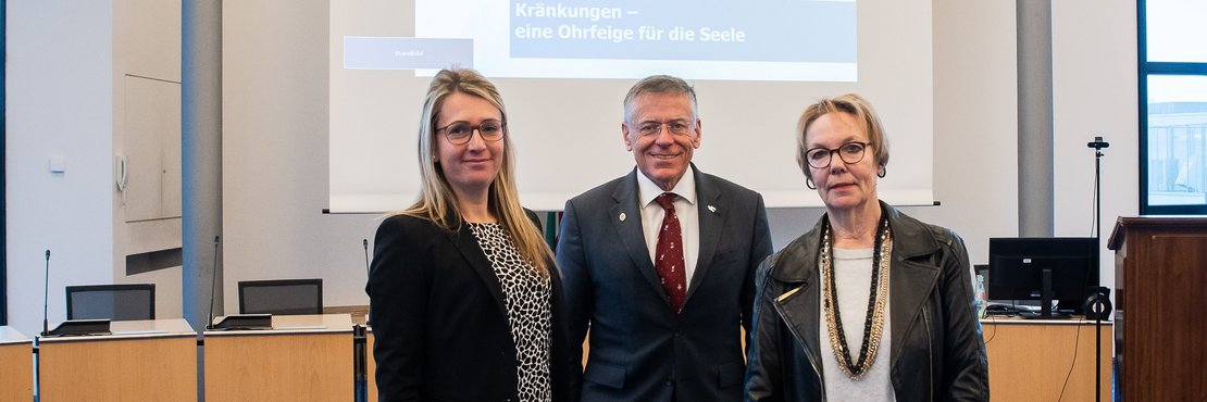 Sabine Raecher, Landrat Hans-Jürgen Petrauschke und Referentin Dr. Bärbel Wardetzki stehen in einem Raum vor einer Leinwand