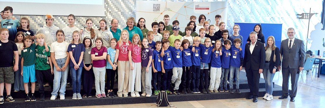 Eine große Gruppe bestehend aus ca. 50 Kindern und einigen Erwachsenen stehe in einer Aula auf einer Bühne. Auf dem Boden sind zwei schmale längliche Parcoure für Roboter abgebildet.