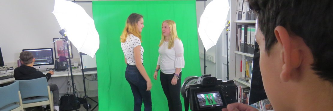Eine männliche Person mit Kamera filmt zwei weibliche Personen vor einem grünen Hintergrund