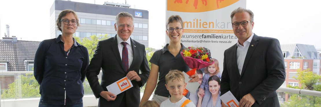 Hans-Jürgen Petrauschke, der Neusser Bürgermeister Reiner Breuer, Marion Klein und Sina Fabri mit ihren beiden Kindern vor einem Plaket der Familienkarte und halten ihre Gewinner in der Hand. Im Hintergrund ist die Stadt Neuss zu sehen.