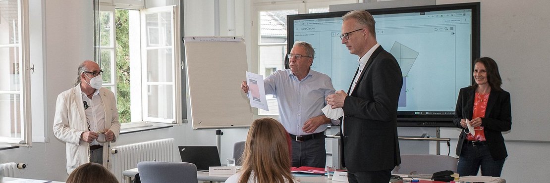 In der Beschreibung benannte Personen stehen in einem Klassenraum. Schüler sitzen an Tischen. Fenster sind geöffnet. Im Hintergrund ein Multimedia-Bildschirm mit mathematischen Zeichnungen. 