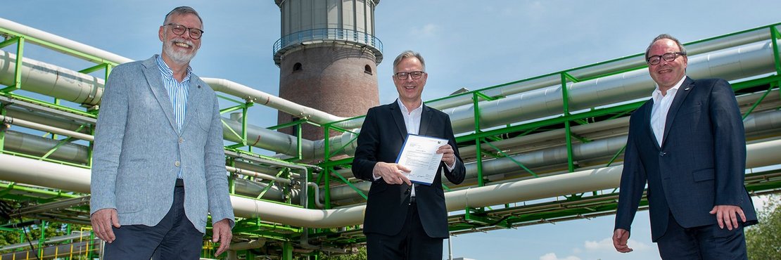 Die drei Männer stehen vor Industrie-Rohrleitungen und einem Turm