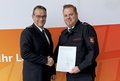 Im Bild sind zwei Männer zu sehen, die sich vor einer orangefarbenen und weißen Wand die Hände schütteln. Der Mann links trägt einen dunklen Anzug, ein weißes Hemd und eine dunkle Krawatte. Seine Brille hat einen dunklen Rahmen und er hat kurze, dunkelbraune Haare. Er wirkt seriös und professionell. Der Mann rechts trägt eine schwarze Feuerwehruniform mit dem Aufdruck „Feuerwehr“ auf der Brusttasche und einem Abzeichen auf dem Ärmel. Seine Haare sind kurz und rotblond. Er hält eine weiße Urkunde mit dem Text „rhein kreis neuss Urkunde“ in den Händen. Die Stimmung ist freundlich und respektvoll, was durch den Händedruck der beiden Männer vermittelt wird. Der Boden ist hellgrau.