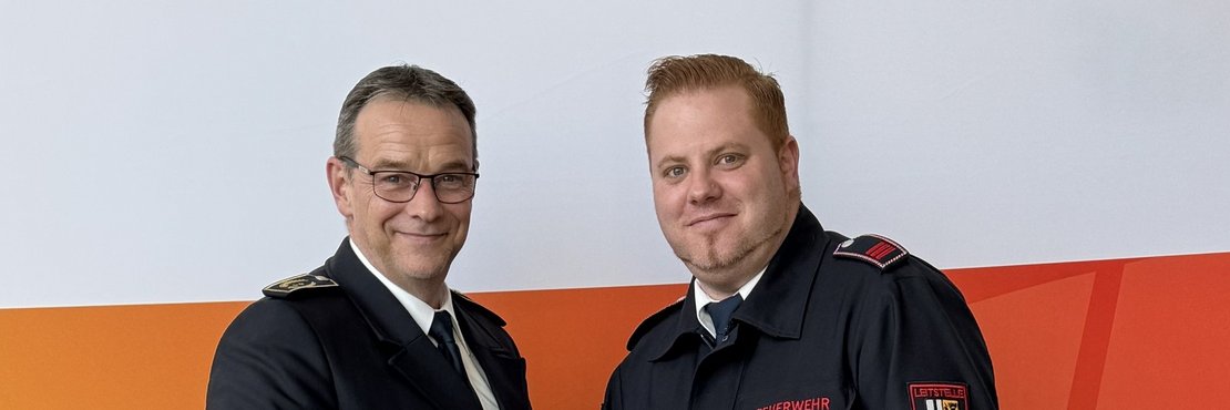 Im Bild sind zwei Männer zu sehen, die sich vor einer orangefarbenen und weißen Wand die Hände schütteln. Der Mann links trägt einen dunklen Anzug, ein weißes Hemd und eine dunkle Krawatte. Seine Brille hat einen dunklen Rahmen und er hat kurze, dunkelbraune Haare. Er wirkt seriös und professionell. Der Mann rechts trägt eine schwarze Feuerwehruniform mit dem Aufdruck „Feuerwehr“ auf der Brusttasche und einem Abzeichen auf dem Ärmel. Seine Haare sind kurz und rotblond. Er hält eine weiße Urkunde mit dem Text „rhein kreis neuss Urkunde“ in den Händen. Die Stimmung ist freundlich und respektvoll, was durch den Händedruck der beiden Männer vermittelt wird. Der Boden ist hellgrau.