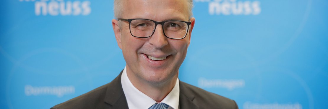 Kreisdirektor Dirk Brügge lächelnd vor blauer Fotowand mit Logos Rhein-Kreis Neuss