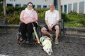 Frau im Rollstuhl mit Assistenzhund. Daneben sitzt ein Mann.