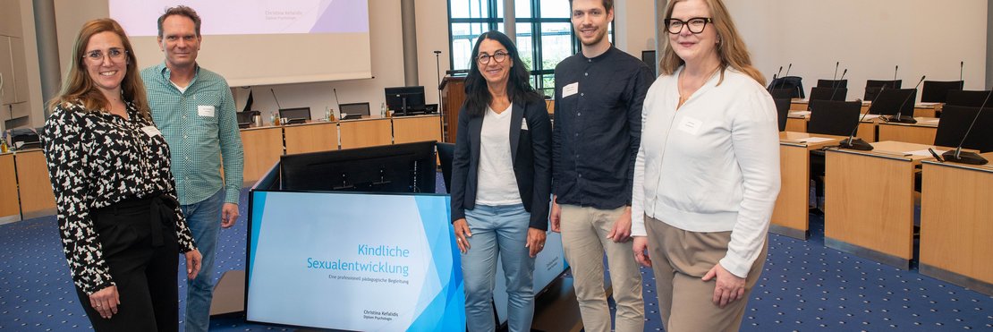 Links und rechts von einem Bildschirm mit der Aufschrift "Kindliche Sexualentwicklung" stehen drei Frauen und zwei Männer.