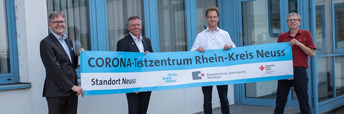 Die genannten Personen stehen vor einem Gebäude. In Ihren Händen halten sie gemeinsam ein langes Banner. Auf diesem steht geschrieben: CORONA-Testzentrum Rhein-Kreis Neuss; Standaort Neuss, Logos sind zu sehen: Rhein-Kreis Neuss, Kassenärztliche Vereinigung Nordrhein und Deutsches Rotes Kreuz