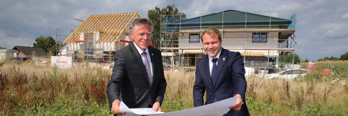 Landrat Petrauschke und Bürgermeister Dr. Mertens stehen auf einer Wiese und halten einen Bauplan. Im Hintergrund die Baustelle des Mehrfamilienhauses.