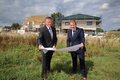 Landrat Petrauschke und Bürgermeister Dr. Mertens stehen auf einer Wiese und halten einen Bauplan. Im Hintergrund die Baustelle des Mehrfamilienhauses.