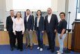 Das Foto zeigt sechs Personen, die vor einem Podium in einem Konferenzraum stehen und für ein Gruppenfoto posieren.