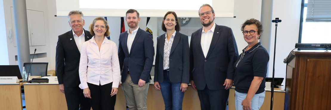 Das Foto zeigt sechs Personen, die vor einem Podium in einem Konferenzraum stehen und für ein Gruppenfoto posieren.