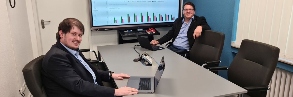 Tobias Schellhorn und Jürgen Brings an Laptops vor einem großen Bildschirm