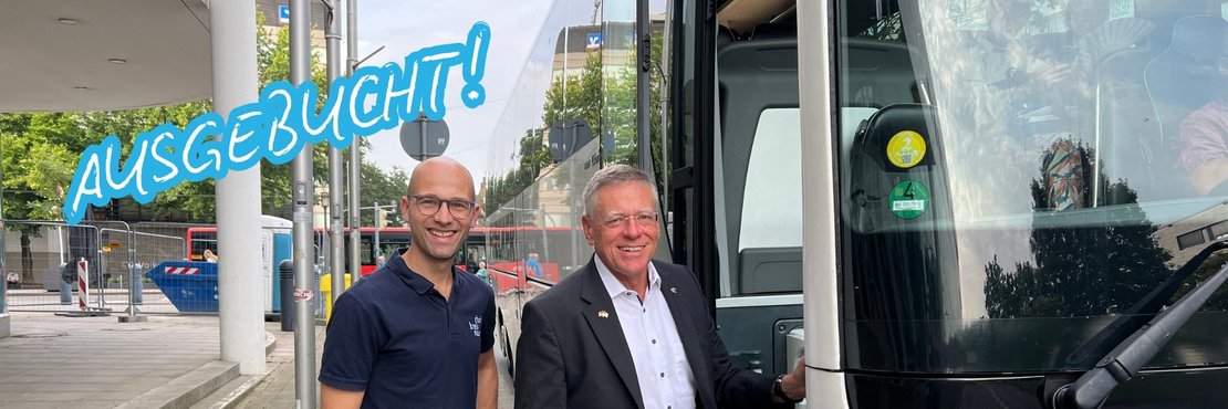 Zwei männliche Personen stehen an der Eingangstüre eine weißen Reisebusses und lächeln.
