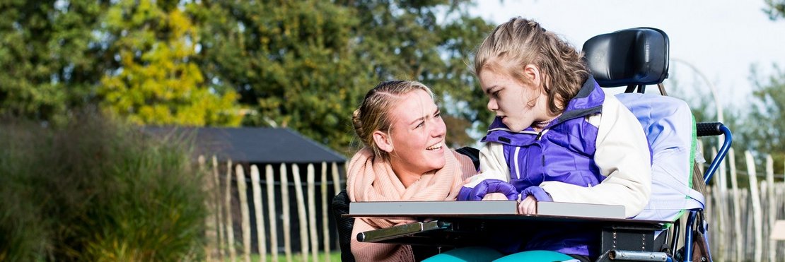 Das Bild zeigt eine junge Frau, die sich liebevoll um ein Mädchen im Rollstuhl kümmert. Beide sitzen auf einem Rasen mit Herbstlaub im Hintergrund. Die Frau lächelt das Mädchen an, und die Szene strahlt Wärme und Zuneigung aus.  Es handelt sich vermutlich um eine fürsorgliche Betreuungssituation. Die Umgebung ist friedlich und natürlich.
