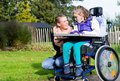 Das Bild zeigt eine junge Frau, die sich liebevoll um ein Mädchen im Rollstuhl kümmert. Beide sitzen auf einem Rasen mit Herbstlaub im Hintergrund. Die Frau lächelt das Mädchen an, und die Szene strahlt Wärme und Zuneigung aus.  Es handelt sich vermutlich um eine fürsorgliche Betreuungssituation. Die Umgebung ist friedlich und natürlich.