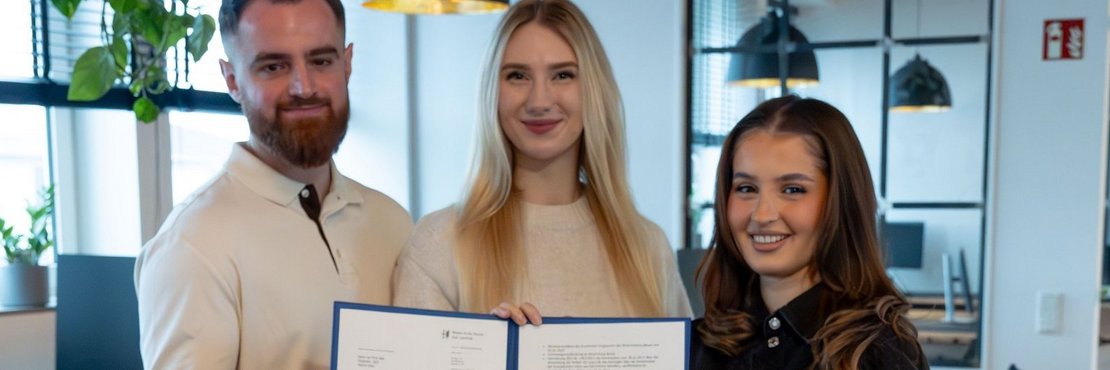 Das Foto zeigt drei junge Erwachsene in einem modernen Büro. Zwei Frauen und ein Mann halten ein gemeinsames Zertifikat oder eine Urkunde in den Händen. Sie lächeln in die Kamera. Der Hintergrund ist unscharf, aber man sieht Schreibtische und moderne Büromöbel.  Das Bild wirkt positiv und vermittelt den Eindruck von Erfolg und Zusammenarbeit.