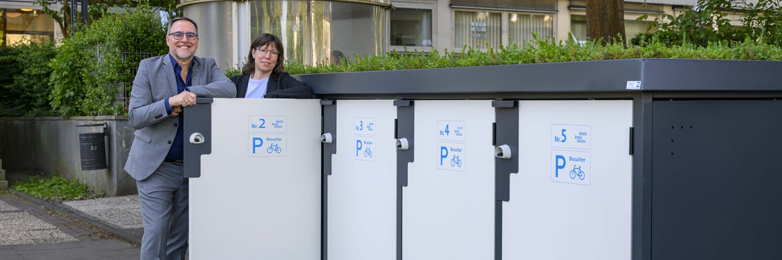 Eine Frau und ein Mann stehen an grau-weißen Fahrradboxen.