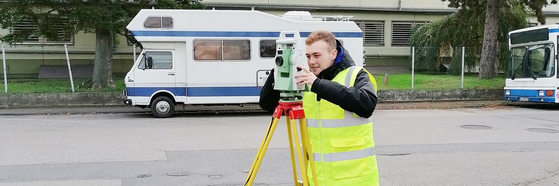 Ein junger Mann in gelber Warnwester blickt durch ein Messgerät. Dieses steht auf einem Dreibein-Stativ auf einer Straße.