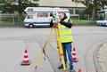 Ein junger Mann in gelber Warnwester blickt durch ein Messgerät. Dieses steht auf einem Dreibein-Stativ auf einer Straße.