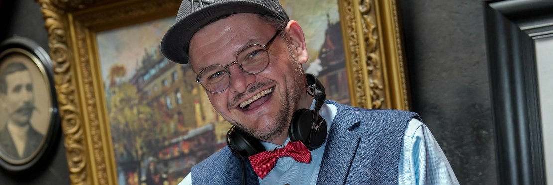 Das Bild zeigt einen Mann mittleren Alters, der hinter einem DJ-Pult steht und Musik auflegt. Er trägt eine Cappy, ein hellblaues Hemd, eine Weste und eine Fliege. Im Hintergrund hängen gerahmte Bilder an der Wand.