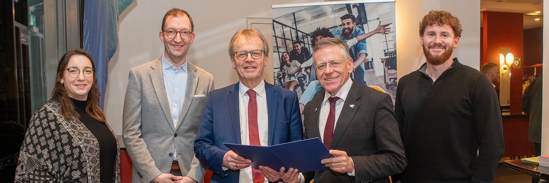 Fünf Personen stehen vor einem großen Banner. Die beiden Personen in der Mitte halten eine blaue Urkunde in den Händen. Die drei anderen Personen stehen neben diesen und lächeln in die Kamera. Die Person ganz links ist eine Frau mit langen braunen Haaren. Neben ihr steht ein Mann in einem hellgrauen Anzug. Der Mann in der Mitte trägt einen dunkelblauen Anzug mit roter Krawatte. Neben ihm steht ein weiterer Mann in einem dunklen Anzug mit roter Krawatte. Ganz rechts steht ein Mann mit kurzem, lockigem, braunen Haar in einem dunklen Pullover und grauen Hosen. Das Banner im Hintergrund zeigt Fotos von jungen, fröhlichen Menschen in einer modernen Arbeitsumgebung.