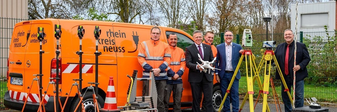 Das Bild zeigt sechs Männer vor einem leuchtend orangefarbenen Lieferwagen. Auf der Seite des Lieferwagens befindet sich ein Logo mit der Aufschrift „Rhein Kreis Neuss“ und einem Stecker-Symbol. Ein Mann steht in der Mitte des Bildes und hält eine weiße Drohne. Vor dem Lieferwagen stehen mehrere Vermessungsgeräte mit Stativen, darunter zwei Totalstationen und ein Nivelliergerät. Ein orangefarbener Verkehrskegel steht ebenfalls im Vordergrund. Im Hintergrund sind Bäume und ein Teil eines Gebäudes zu sehen.