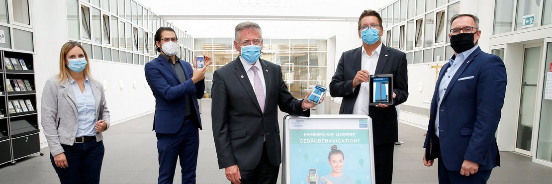 Landrat Hans-Jürgen Petrauschke steht mit einer Frau und frei Herren im Lichthof des Kreishauses Neuss. Landrat Petrauschke und Digitalisierungsmanager Brings halten ein Smartphone bzw. ein Tablet auf dem die Navigatoions-App zu sehen ist.  Mittig auf dem Boden steht ein Plakat mit der Überschrift "Kennen Sie unsere Gebäude-Navigation?" und Hinweisen zur App.