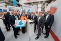 Das Bild zeigt eine Gruppe von etwa 14 Personen, hauptsächlich Männer in Anzügen, die vor einem Informationsstand auf einer Messe stehen.  Im Vordergrund hält eine Frau eine Karte des Rhein-Kreises Neuss.  Die Männer im Hintergrund tragen ebenfalls Anzüge. Die Schilder weisen auf die Kreise Wesel und Rhein-Kreis Neuss hin. Es scheint sich um eine Veranstaltung zum Thema Wirtschaftsförderung zu handeln. Die Atmosphäre wirkt formell und repräsentativ.