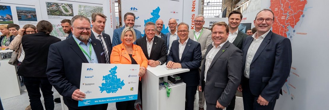 Das Bild zeigt eine Gruppe von etwa 14 Personen, hauptsächlich Männer in Anzügen, die vor einem Informationsstand auf einer Messe stehen.  Im Vordergrund hält eine Frau eine Karte des Rhein-Kreises Neuss.  Die Männer im Hintergrund tragen ebenfalls Anzüge. Die Schilder weisen auf die Kreise Wesel und Rhein-Kreis Neuss hin. Es scheint sich um eine Veranstaltung zum Thema Wirtschaftsförderung zu handeln. Die Atmosphäre wirkt formell und repräsentativ.