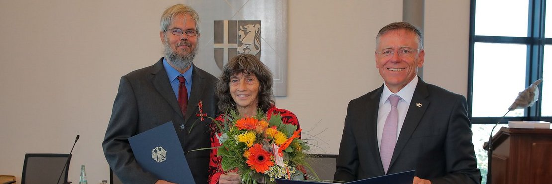Landrat Hans-Jürgen Petrauschke Regina Hoppe-Kresse (mit Blumen) und Martin Kresse, ausgezeichnet mit dem Bundesverdienstkreuz,  stehen lächelnd nebeneinander. 