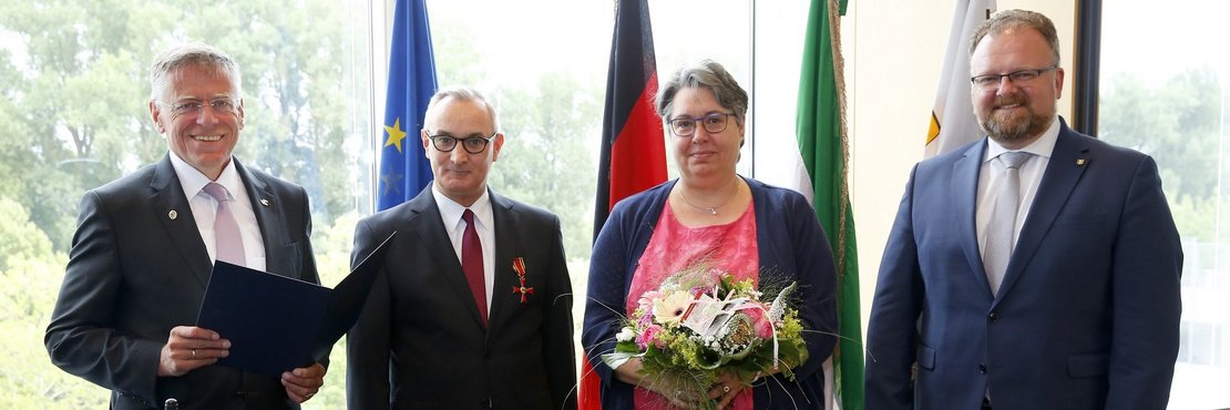 Landrat Hans-Jürgen Petrauschke, Udo Bartsch, Karin Bartsch und Bürgermeister Marc Venten. Frau Bartsch hält einen Strauß Blumen. Im Hintergrund sieht man verschiedene Fahnen. 