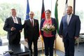Landrat Hans-Jürgen Petrauschke, Udo Bartsch, Karin Bartsch und Bürgermeister Marc Venten. Frau Bartsch hält einen Strauß Blumen. Im Hintergrund sieht man verschiedene Fahnen. 