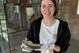 Das Bild zeigt eine junge Frau mit Brille, die in einem Museum vor einer Vitrine steht.  Sie trägt ein schwarzes Sweatshirt und ein weißes T-Shirt und hält mit weißen Baumwollhandschuhen eine silberne Schale mit einer darauf befindlichen Skulptur. Die Schale scheint ein antikes oder kunsthandwerkliches Stück zu sein. Im Hintergrund sind weitere Ausstellungsstücke in der Vitrine zu sehen.