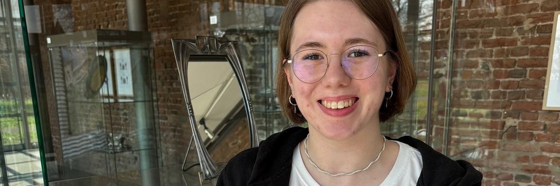 Das Bild zeigt eine junge Frau mit Brille, die in einem Museum vor einer Vitrine steht.  Sie trägt ein schwarzes Sweatshirt und ein weißes T-Shirt und hält mit weißen Baumwollhandschuhen eine silberne Schale mit einer darauf befindlichen Skulptur. Die Schale scheint ein antikes oder kunsthandwerkliches Stück zu sein. Im Hintergrund sind weitere Ausstellungsstücke in der Vitrine zu sehen.