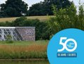 Das Bild zeigt die Langen Foundation im Museum Insel Hombroich bei Sonnenuntergang. Im Vordergrund befindet sich ein ruhiger Wasserbecken, das die moderne Architektur des Museums und die umliegende Landschaft spiegelt. Ein Teil des Museumsgebäudes, das aus Beton und Glas besteht, ist sichtbar.  Im Hintergrund ist eine Betonmauer zu sehen, vor der eine Skulptur steht.  Rechts unten befindet sich ein blaues Logo mit der Aufschrift "50" und dem Hinweis auf ein Jubiläum ("50 Jahre | 50 Orte"). Das Bild wirkt ruhig und atmosphärisch.