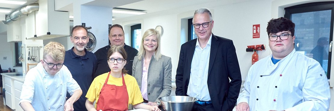 Personengruppe mit 7 Menschen stehen vor einer Küchentheke mit Lebensmitteln darauf.