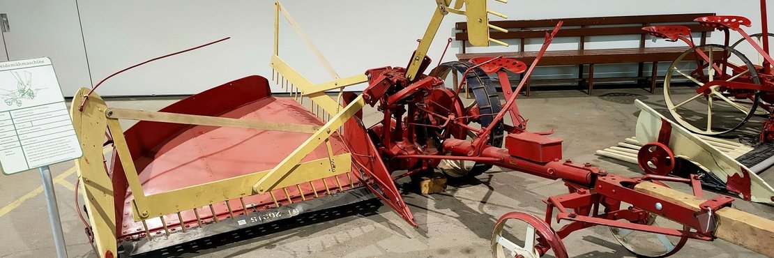 Das Bild zeigt eine alte Heumachmaschine (oder eine ähnliche landwirtschaftliche Maschine) in einem Museum oder einer ähnlichen Ausstellungshalle. Die Maschine ist rot und gelb lackiert und steht auf dem Boden. Es gibt auch einige andere landwirtschaftliche Geräte im Hintergrund zu sehen sowie ein Hinweisschild mit einem technischen Diagramm der Maschine.
