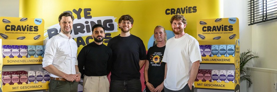 Das Bild zeigt fünf Personen, die vor einem aufgestellten Display mit Verpackungen von „Cravies“ Sonnenblumenkernen stehen.  Es scheint sich um eine Produktvorstellung oder eine Marketingveranstaltung zu handeln.  Die Personen wirken entspannt und lächeln in die Kamera. Das Hintergrundbanner bewirbt die Produkte mit dem Slogan „Bye Boring Snacks“.  Die Personen stehen auf einem roten Teppich.  Zwei der Personen sind Männer in Hemd beziehungsweise Pullover, eine Frau trägt ein T-Shirt mit einem Sonnenblumenmotiv, und zwei weitere Männer tragen jeweils ein T-Shirt.
