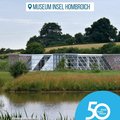 Das Bild zeigt die Cafeteria des Museums Insel Hombroich, ein modernes Gebäude aus Glas und Stein, das sich harmonisch in die umliegende Naturlandschaft einfügt. Im Vordergrund ist ein ruhiger See mit Schilfbewuchs zu sehen, im Hintergrund erstreckt sich eine grüne Hügellandschaft.  Ein blauer Kreis mit der Aufschrift "50 Jahre | 50 Orte" und dem Logo des Rhein-Kreises Neuss rundet das Bild ab.  Das Gesamtbild vermittelt Ruhe, Naturverbundenheit und die Ästhetik des Museums. © Museum Insel Hombroich / Thomas Riehle, Erwin Heerich