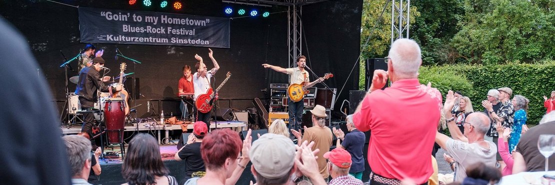 Das Bild zeigt eine Open-Air-Blues-Rock-Konzertveranstaltung namens „Goin’ to my Hometown“ im Kulturzentrum Sinsteden. Eine Band spielt auf einer Bühne vor einem begeisterten Publikum, das klatscht und mit den Händen gestikuliert. Die Stimmung ist ausgelassen und fröhlich. Im Hintergrund sind Bäume und ein Teil des Veranstaltungsortes zu sehen.