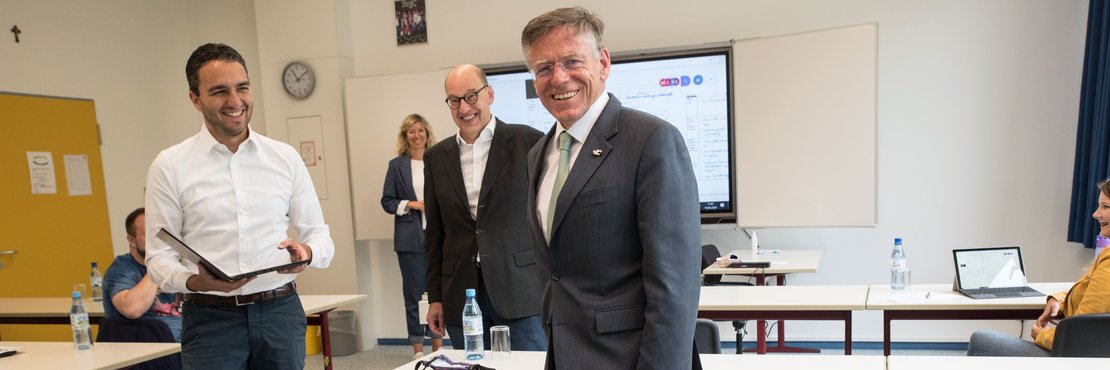 Christopher Drews und Elke Klehm mit Tillmann Lonnes und Landrat Hans-Jürgen Petrauschke stehen mit Abstand in einem Klassenzimmer mit Laptop und großem Bildschirm im Hintergrund