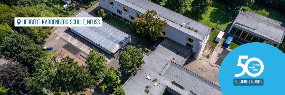 Das Bild zeigt eine Luftaufnahme der Herbert-Karrenberg-Schule in Neuss.  Man sieht die Schulgebäude, den Schulhof mit Spielgeräten und einen Bereich, der möglicherweise im Umbau oder in der Renovierung ist.  Im Vordergrund befindet sich ein Logo, das auf das 50-jährige Jubiläum des Rhein-Kreises Neuss hinweist. Das Bild vermittelt einen Eindruck vom Schulgelände und verbindet dies mit dem Jubiläum des Kreises.