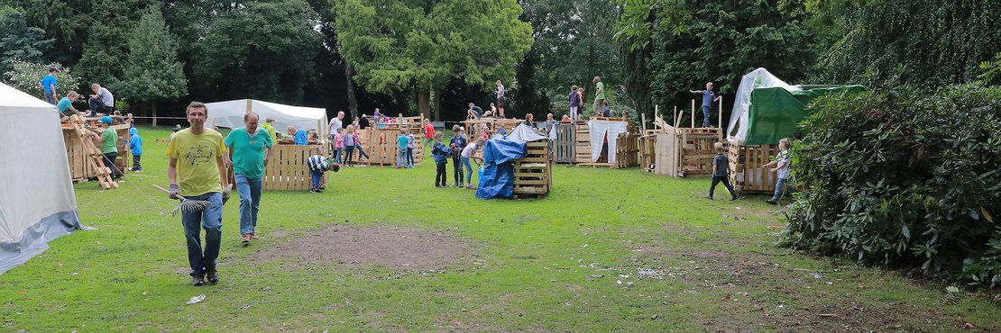 Auf dem Bild ist eine Gruppe von Kindern und Erwachsenen zu sehen, die in einem Waldstück aus Paletten verschiedene Spiel- und Unterstände bauen.  Das Bild zeigt eine lebhafte Szene auf einem grünen Rasen, umgeben von hohen Bäumen eines Waldes. Im Vordergrund steht eine Gruppe von Personen, zwei Erwachsene, die in Richtung Kamera gehen und sich mit einem Rechen und anderen Materialien beschäftigen. Im Hintergrund sind mehrere Kinder und einige Erwachsene beschäftigt, Holzkisten und -paletten zu verschiedenen Strukturen umzubauen, die als kleine Häuser, Plattformen und Türme aussehen. Einige Kinder klettern auf den Konstruktionen herum, andere scheinen an ihrem Bau beteiligt zu sein. Es gibt zwei große, helle Zelte im Hintergrund, die wahrscheinlich als Aufenthaltsräume dienen. Die Atmosphäre ist fröhlich und aktiv, die Stimmung ist spielerisch und kreativ. 