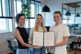 Das Bild zeigt drei Personen, zwei Frauen und einen Mann, die in einem modernen Büro vor einem hellen Holzhintergrund stehen. Sie halten gemeinsam ein Zertifikat oder eine Urkunde in den Händen. Die Frauen tragen Jeans und der Mann beige Hosen. Die Atmosphäre wirkt professionell und positiv.