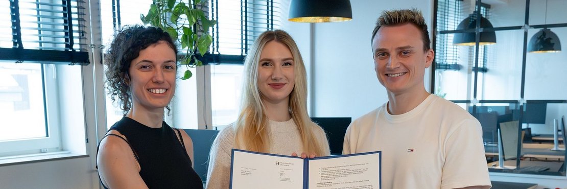 Das Bild zeigt drei Personen, zwei Frauen und einen Mann, die in einem modernen Büro vor einem hellen Holzhintergrund stehen. Sie halten gemeinsam ein Zertifikat oder eine Urkunde in den Händen. Die Frauen tragen Jeans und der Mann beige Hosen. Die Atmosphäre wirkt professionell und positiv.