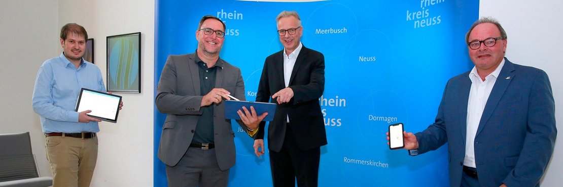 Die vier Männer stehen mit Smartphones und Tablets vor einer blauen Fotowand mit Logos des Rhein-Kreises Neuss