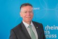 Hans-Jürgen Petrauschke, Landrat des Rhein-Kreis Neuss, lächelnd vor einem hellblauen Hintergrund